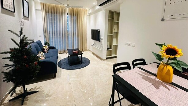 En moderne stue med sofa, et lille juletræ, et spisebord med stole og et stort vindue med gardiner.