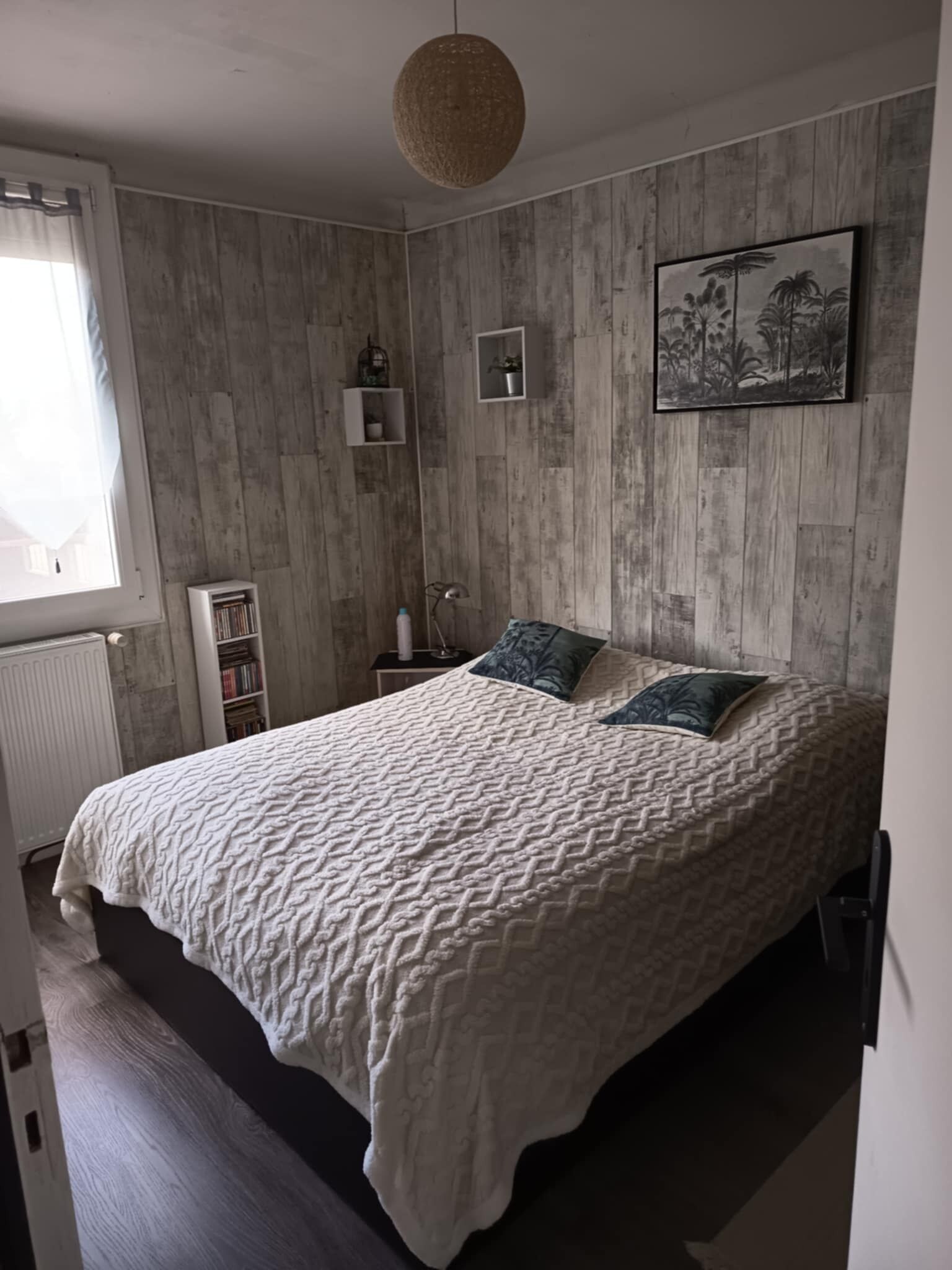 Une chambre avec un grand lit, un panneau mural en bois, une fenêtre avec des rideaux, une table de chevet avec une lampe et un tableau encadré au mur.
