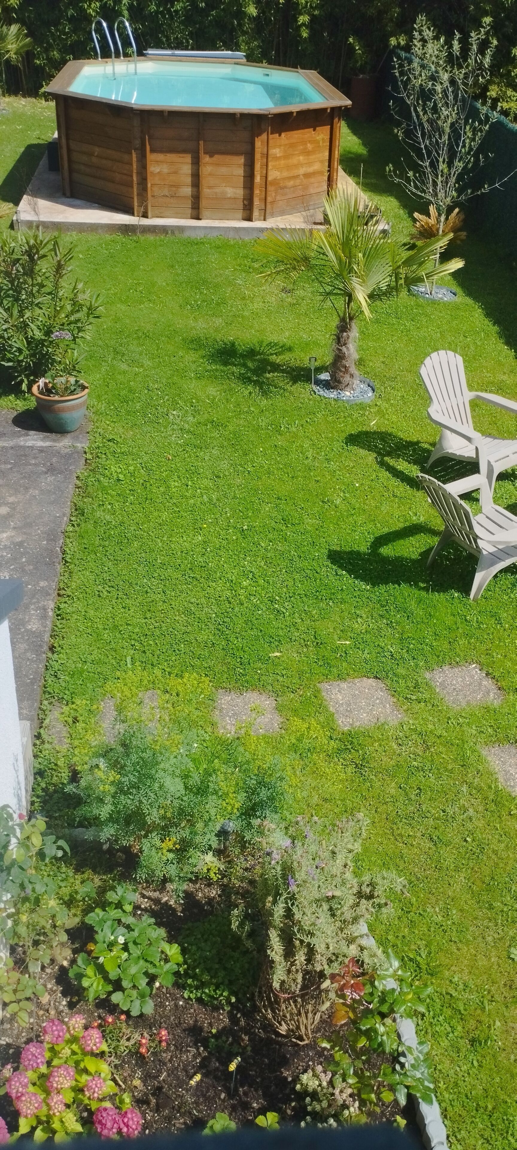 A wooden hot tub with steps, surrounded by a well-maintained lawn, potted plants, and a white Adirondack chair.