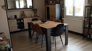 Eine moderne Küche mit Essbereich. Sie zeigt einen großen Holztisch mit Stühlen, einen schwarzen Stuhl und eine Schüssel auf dem Tisch. Ein Fenster mit Vorhängen, ein Heizkörper und eine Pflanze auf dem Fensterbrett sind zu sehen.