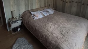 Un dormitorio con pared de madera, una cama con manta estampada, un colgante de luz y una mesita de noche pequeña con una lámpara.