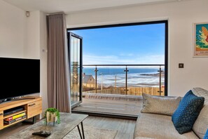 Una sala de estar moderna con un televisor grande, un mueble de madera para entretenimiento y vista a la playa a través de un ventanal.