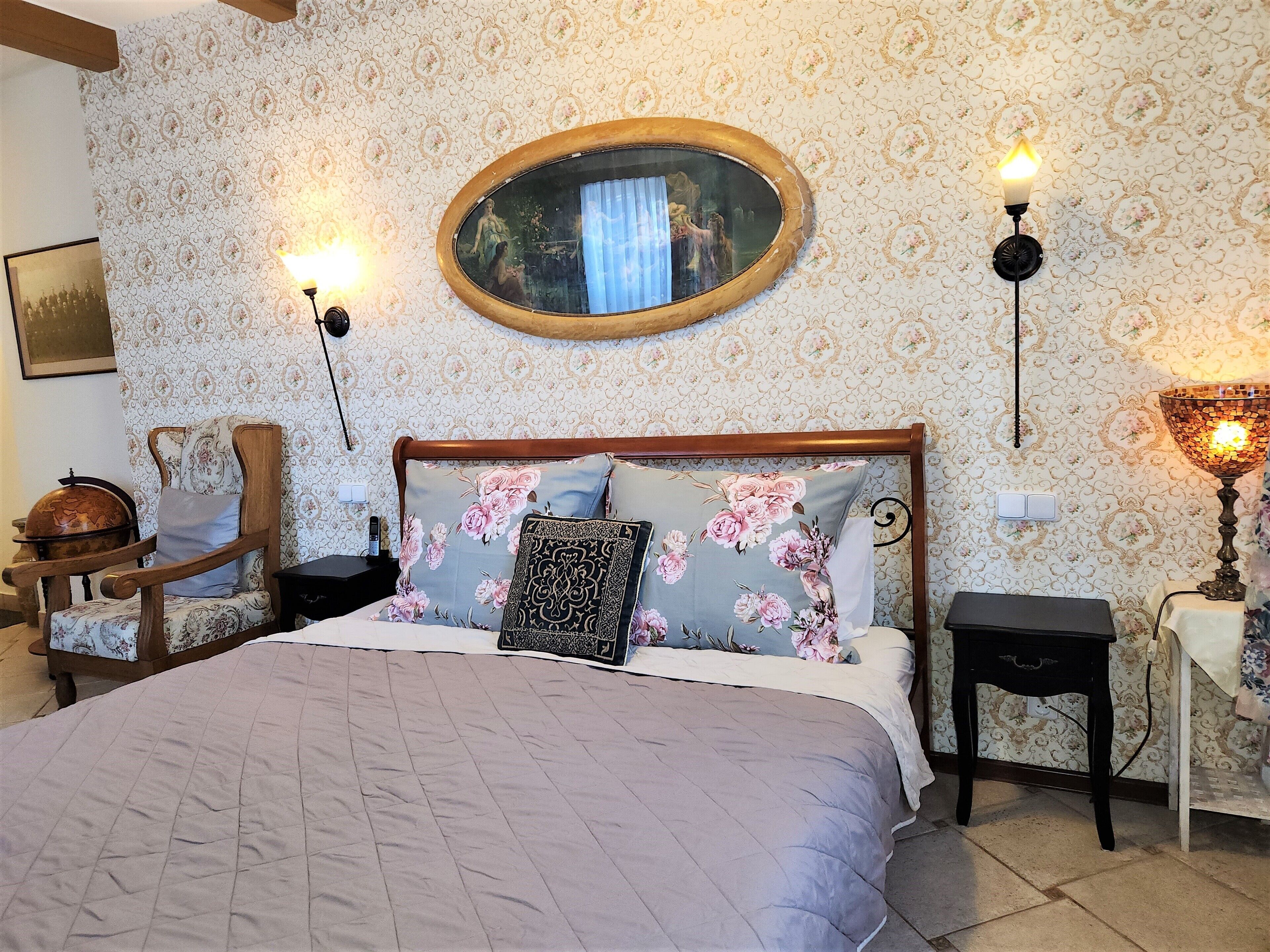 A bedroom with a large bed, floral-patterned pillows, a wooden headboard, a wall-mounted mirror, and two bedside lamps.