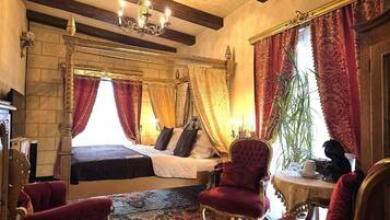 Uma cama com dossel e cortinas vermelhas e douradas, um candelabro e uma poltrona vermelha em um quarto com paredes de pedra e teto de madeira.