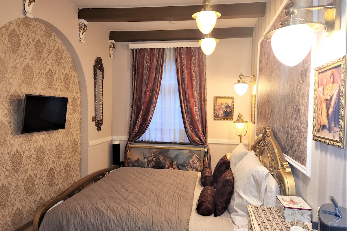 A hotel room with a large bed, a flat-screen TV mounted on the wall, a painting above the bed, and a small table with a cup and saucer.