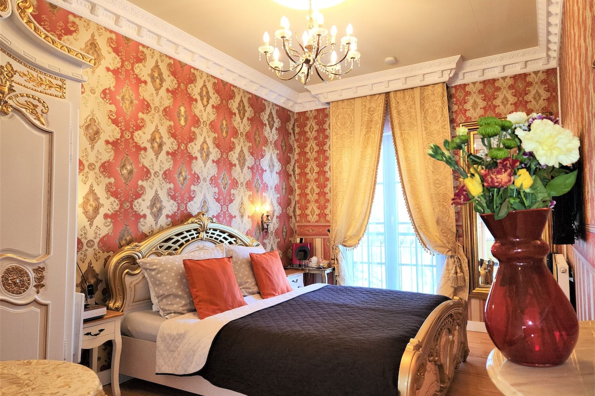 A classic, ornate bedroom with a large bed, a chandelier, and a vase of flowers.