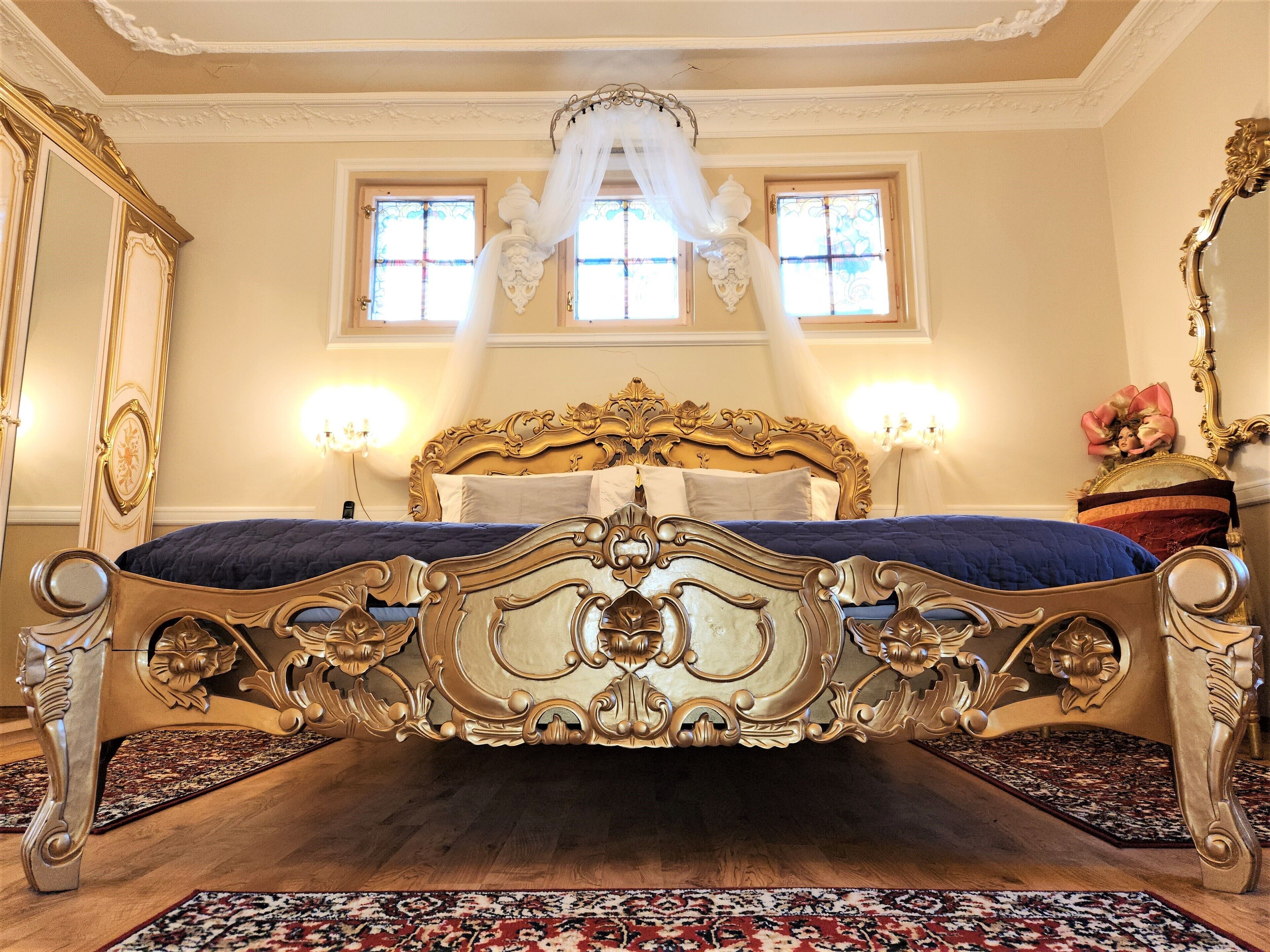 A luxurious bedroom with an ornate four-poster bed, two wall-mounted lamps, and a large mirror with a decorative frame.