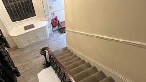 Un escalier avec une rampe en bois et des marches recouvertes de moquette, menant à un palier où se trouvent un banc et un extincteur.