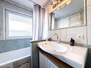 A bathroom with a bathtub, a sink, a mirror, and a window with a curtain. - Staufenhirsch Apartment (Oberstaufen)