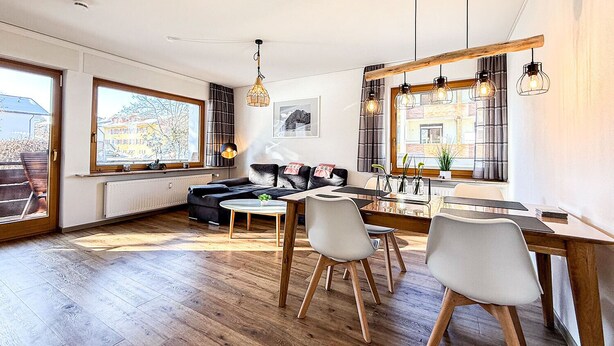 En moderne stue med en sort sofa, et spisebord med stole og store vinduer med udsigt til bygninger.
