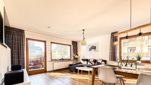 En moderne stue med sofa, spisebord og stole. Store vinduer med gardiner, en radiator og en hængende lampe.