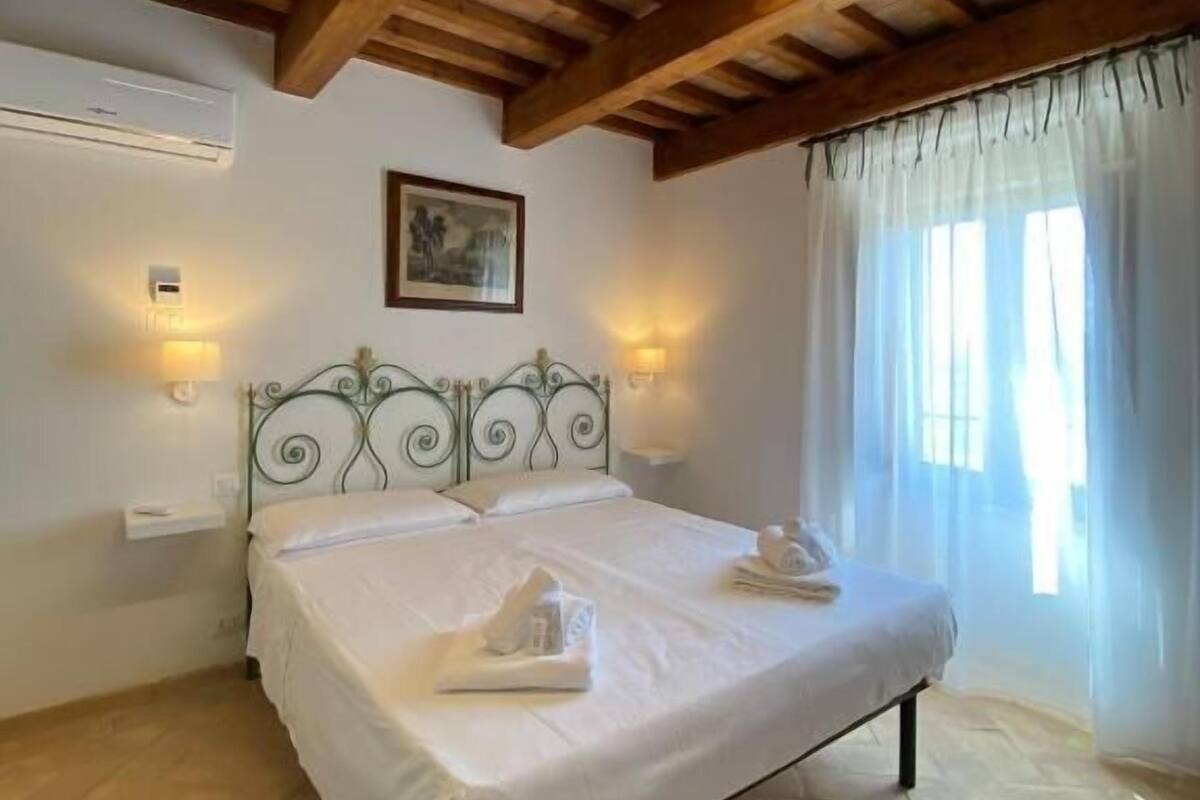 A bedroom with a wooden ceiling, a bed with white linens, two wall-mounted lights, and a window with sheer curtains.