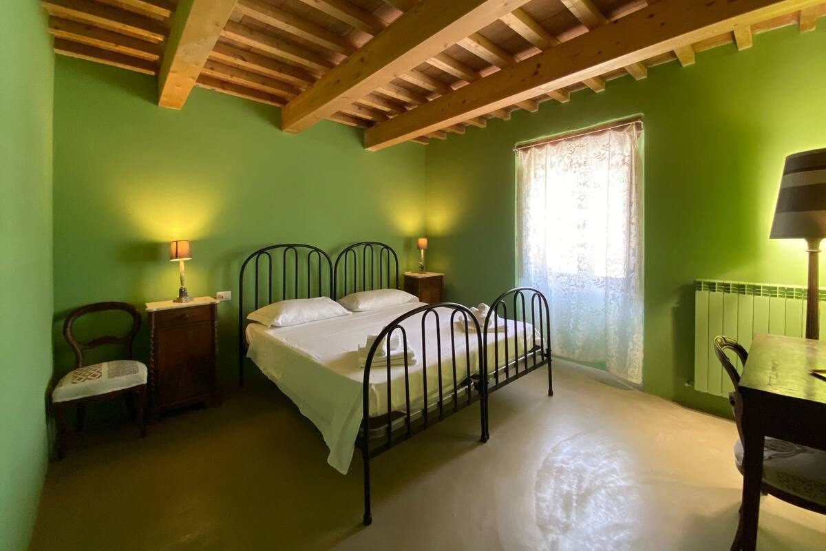A bedroom with a bed, two bedside tables, a chair, a window with curtains, and a wooden ceiling structure.