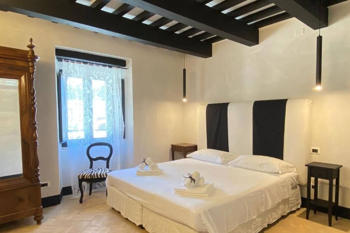A bedroom with a large bed, a wooden wardrobe, a chair, a bedside table, and a window with curtains.