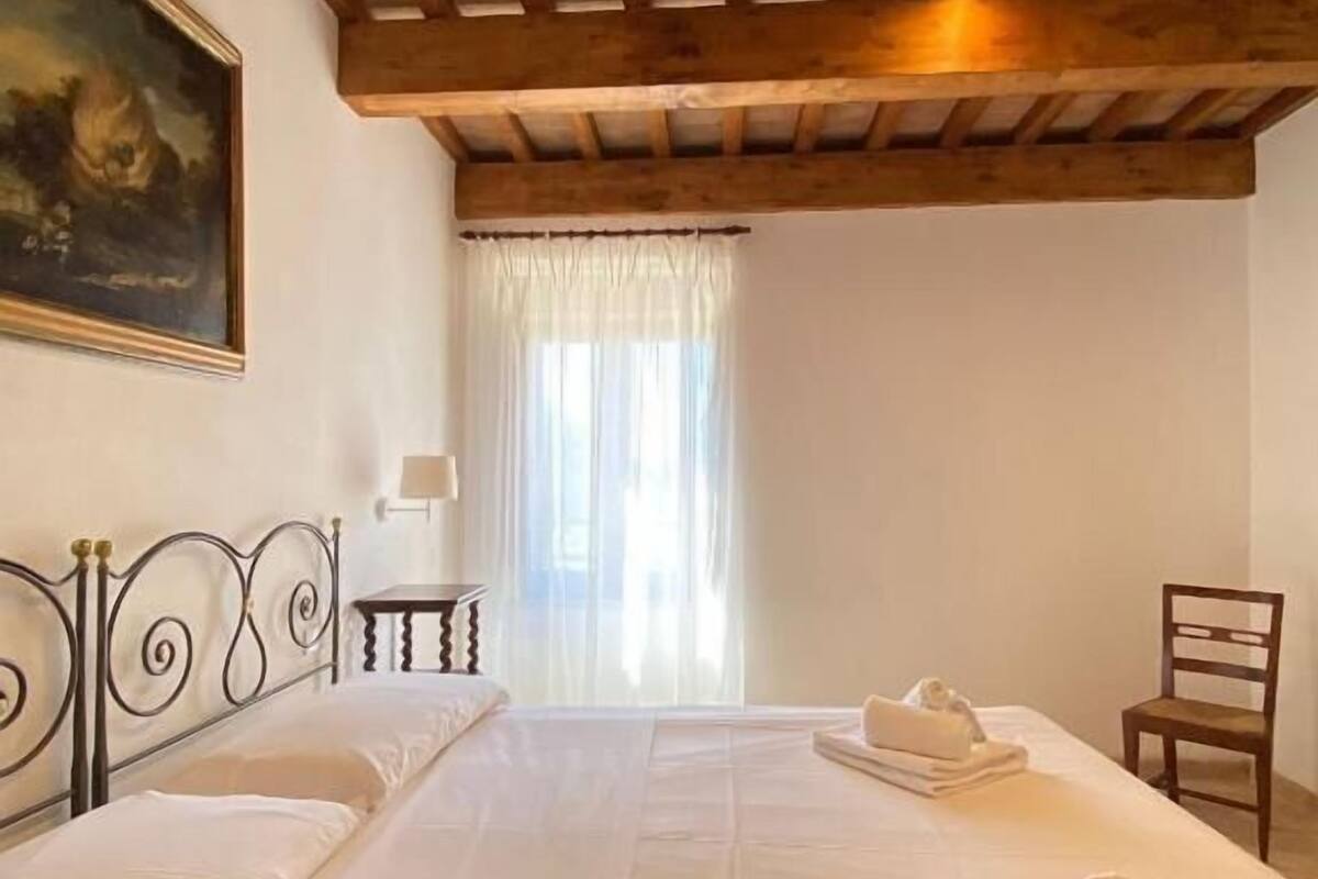 A bedroom with a large bed, wooden ceiling beams, a painting on the wall, a chair, and a window with curtains.