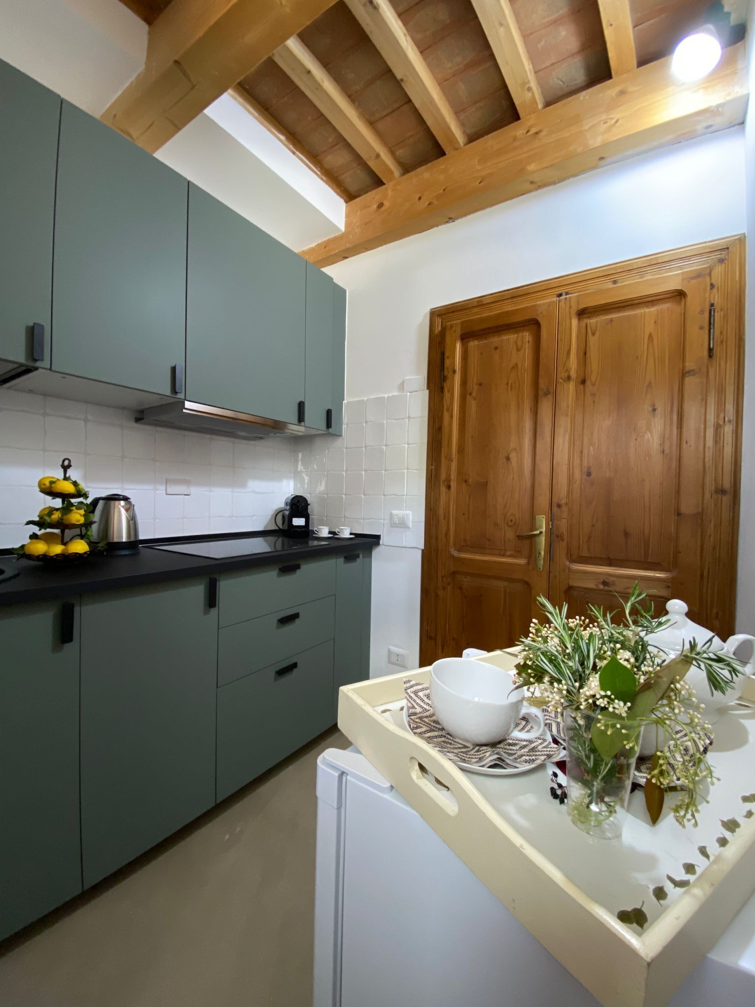 A kitchen with green cabinets, a wooden door, and a countertop with a coffee maker, kettle, and flowers.
