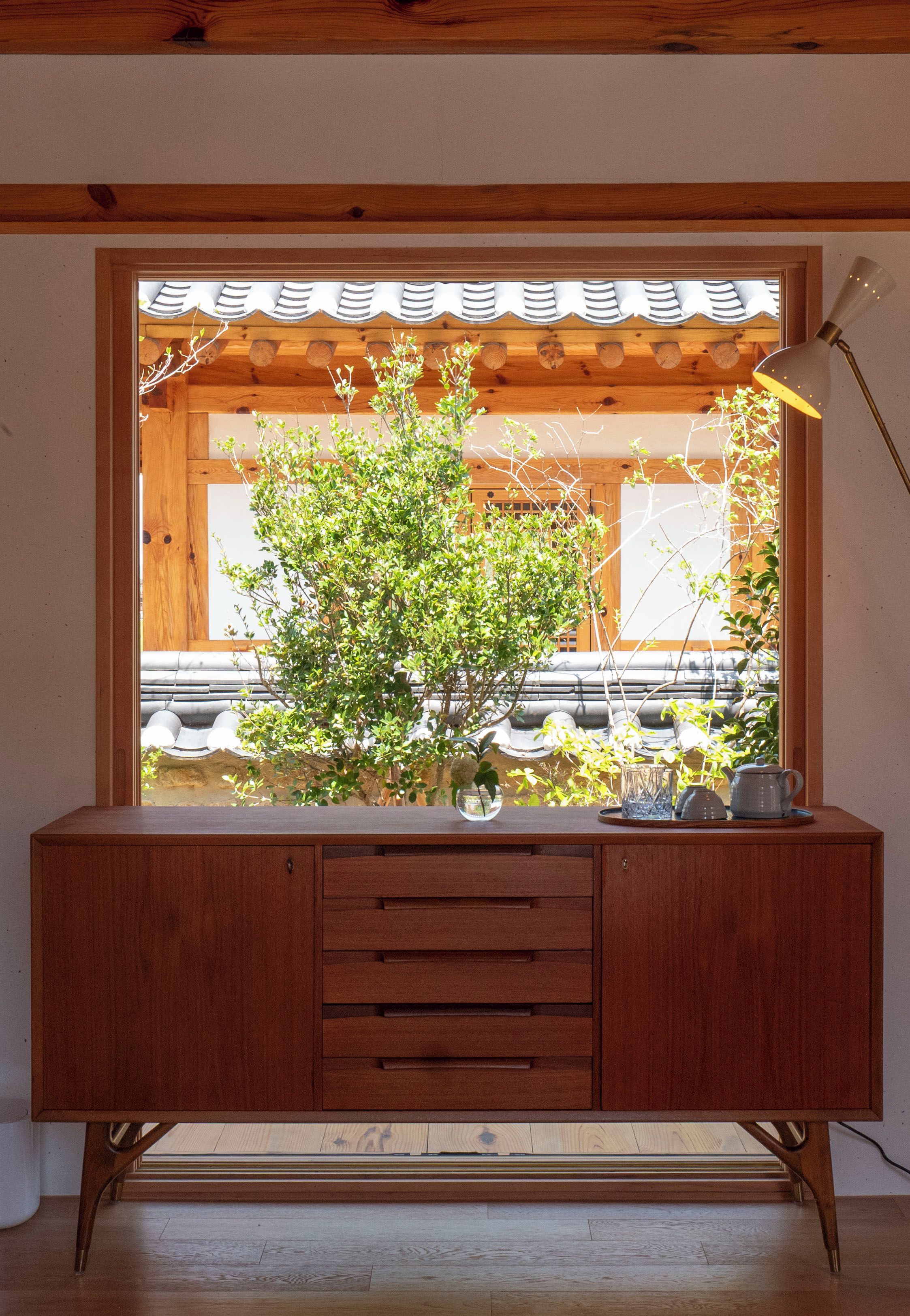 Un mueble de madera con ventana integrada, un jarrón con plantas verdes y una lámpara a la derecha.