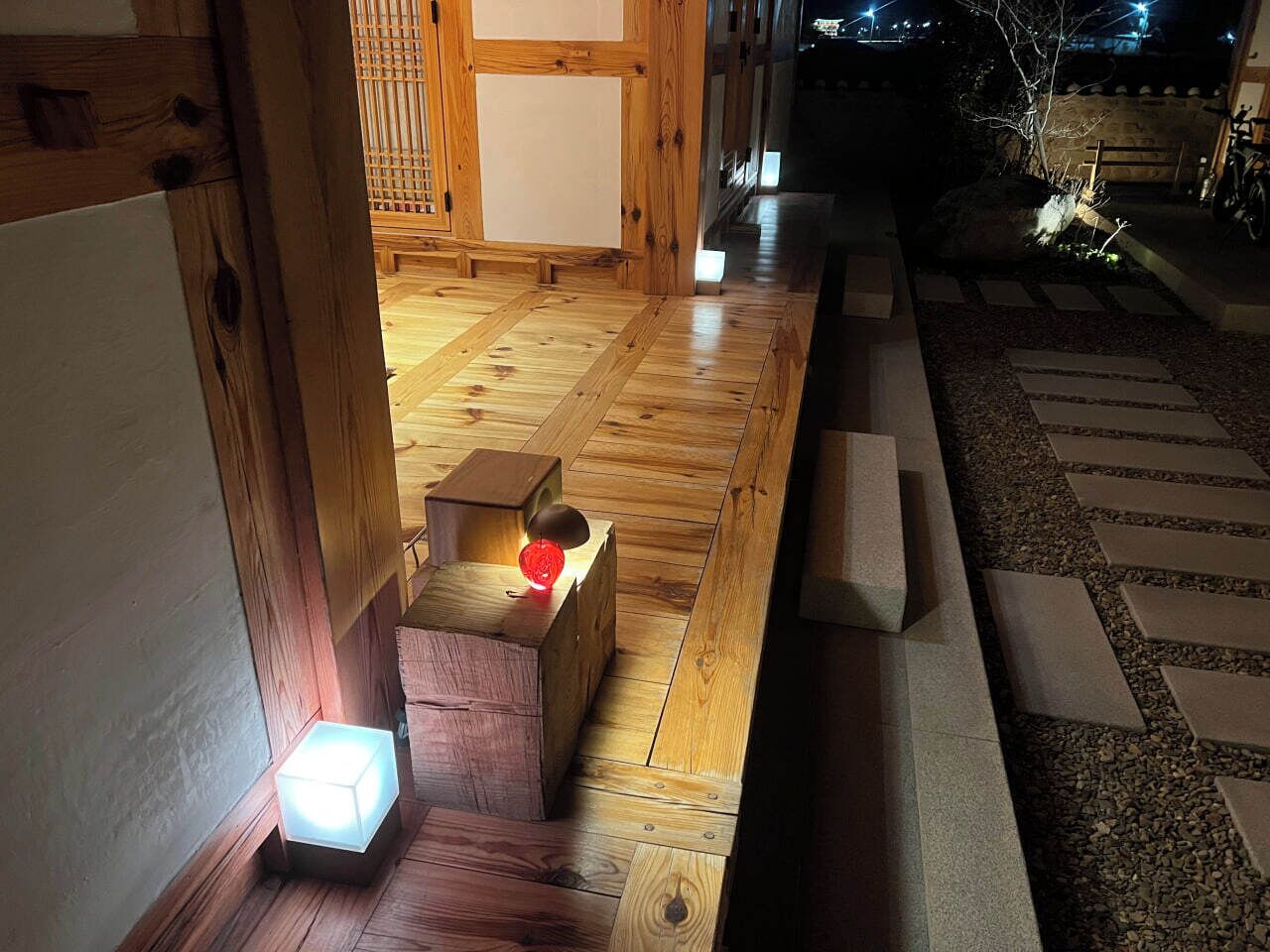 Un intérieur en bois avec un chemin menant à une porte, une petite table sur laquelle se trouve un objet rouge, et une lampe de sol allumée.