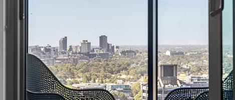 A balcony with two black chairs, a small table, and a view of a cityscape with buildings and trees.