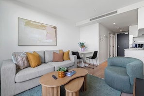 Ein modernes Wohnzimmer mit einem grauen Sofa, einem runden Couchtisch und einem Türkis-Armlehnstuhl. Es gibt einen Essbereich mit weißem Tisch und Stühlen, und im Hintergrund ist eine Küche zu sehen.