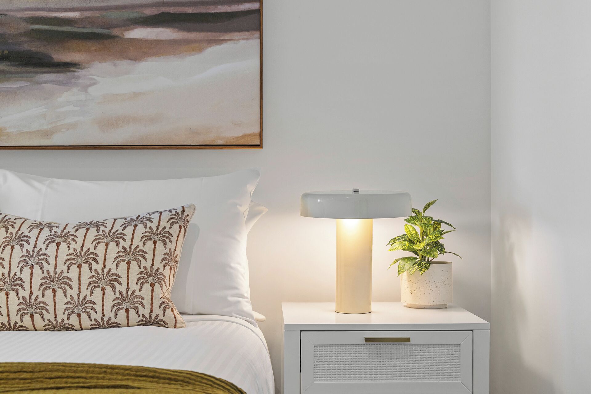 A neatly made bed with a patterned pillow, a bedside table with a lamp and a plant, and a painting on the wall.