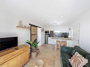 A modern living room with a TV, wooden cabinet, green sofa, and a kitchen in the background. - Appartement 4 personnes, climatisé, Wifi, parking, piscine et vue mer ! (Cavalaire-sur-Mer)