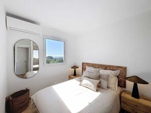 A bedroom with a bed, a bedside table with a lamp, a mirror, a window with a view, and a wicker basket. - Appartement 4 personnes, climatisé, Wifi, parking, piscine et vue mer ! (Cavalaire-sur-Mer)