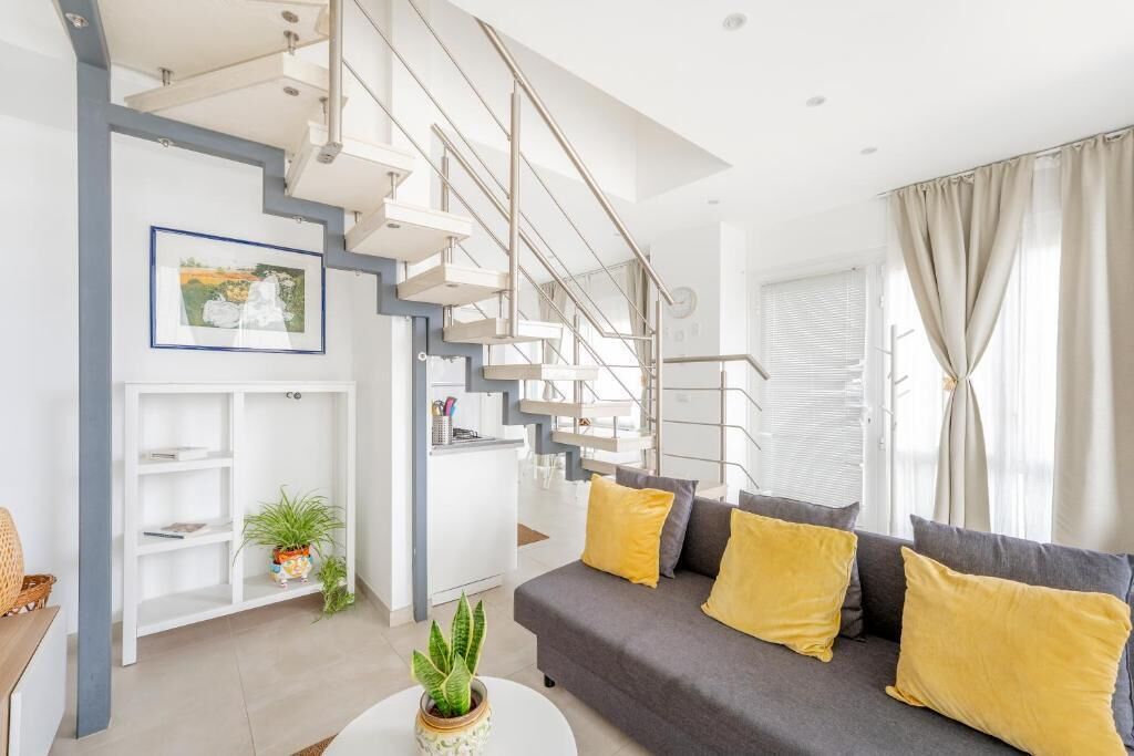 A modern living room with a grey sofa and yellow cushions, a staircase with a white railing, and a bookshelf with a framed picture.