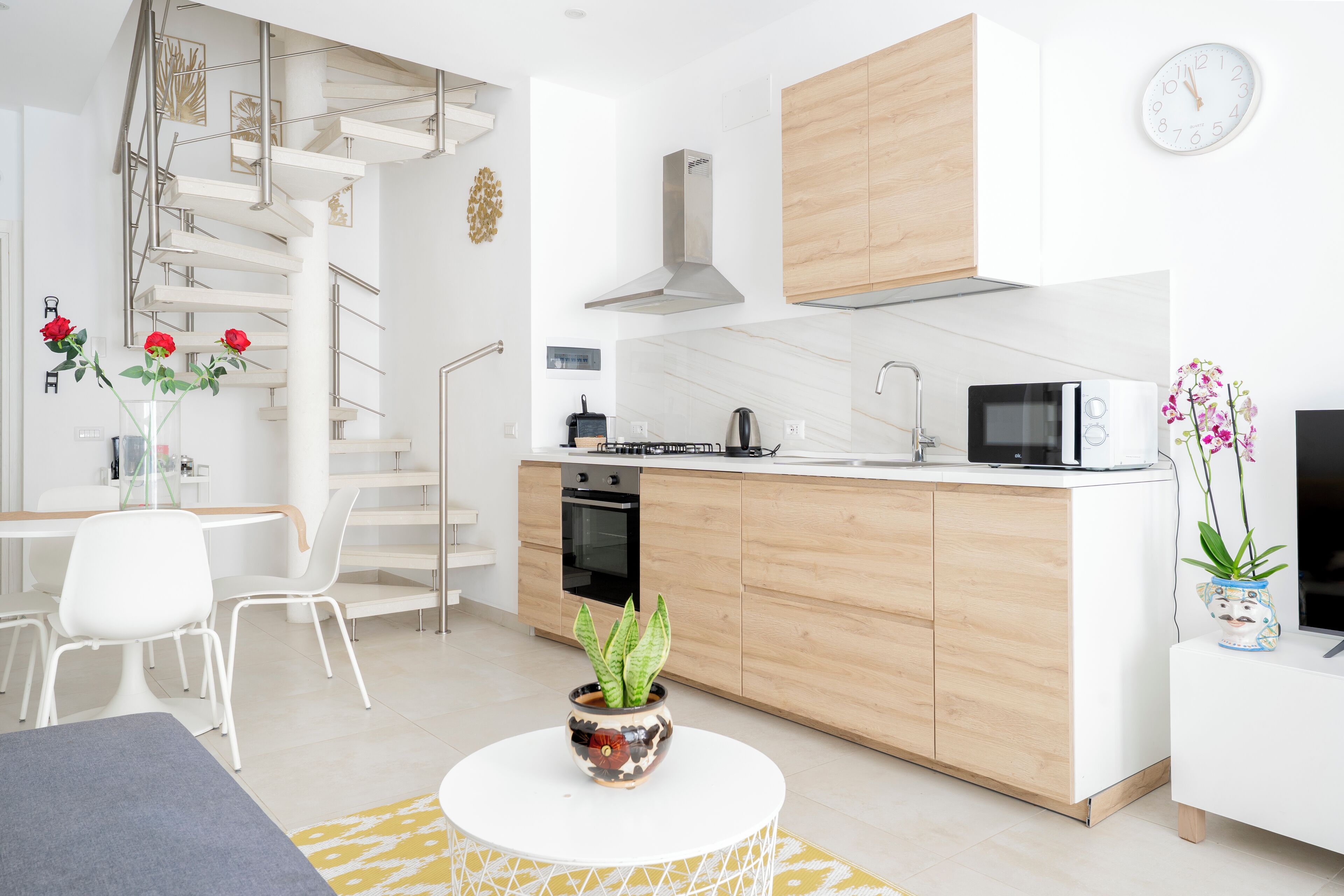 Eine moderne Küche mit weißen Schränken, einem eingebauten Ofen, einer Mikrowelle und einer Edelstahl-Dunstabzugshaube. Ein Essbereich mit weißem Tisch und Stühlen lädt zum Verweilen ein, und eine Treppe führt zu einer oberen Ebene.