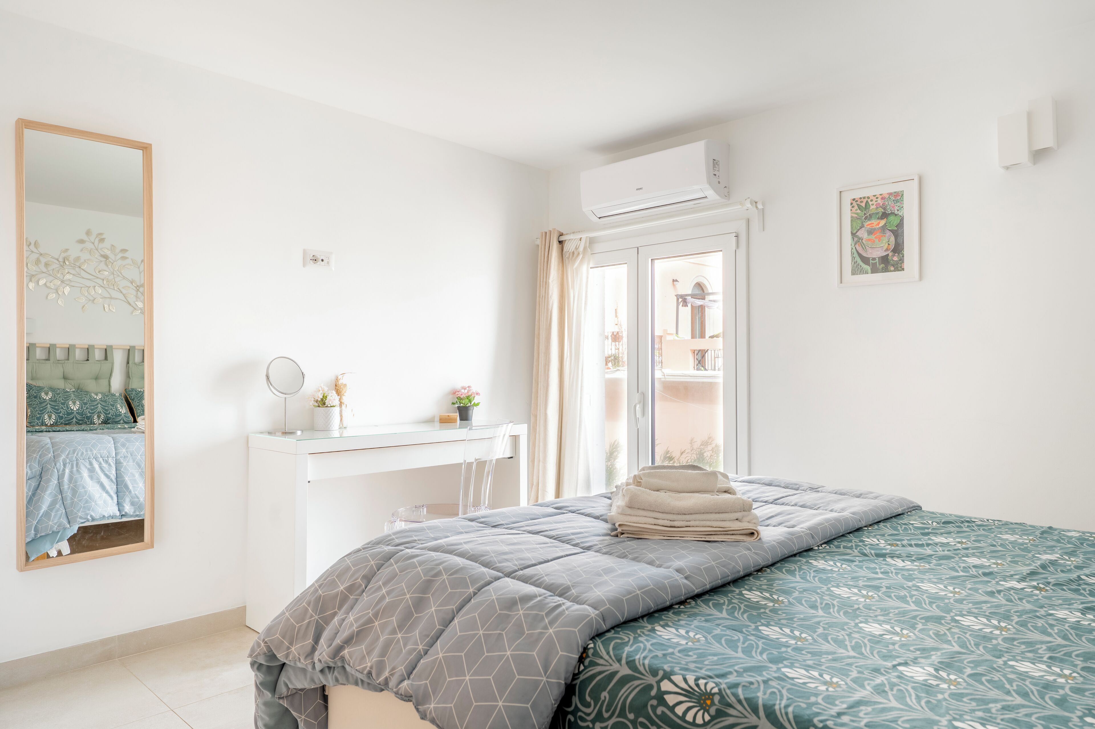A bedroom with a bed, a desk, a mirror, and an air conditioning unit.