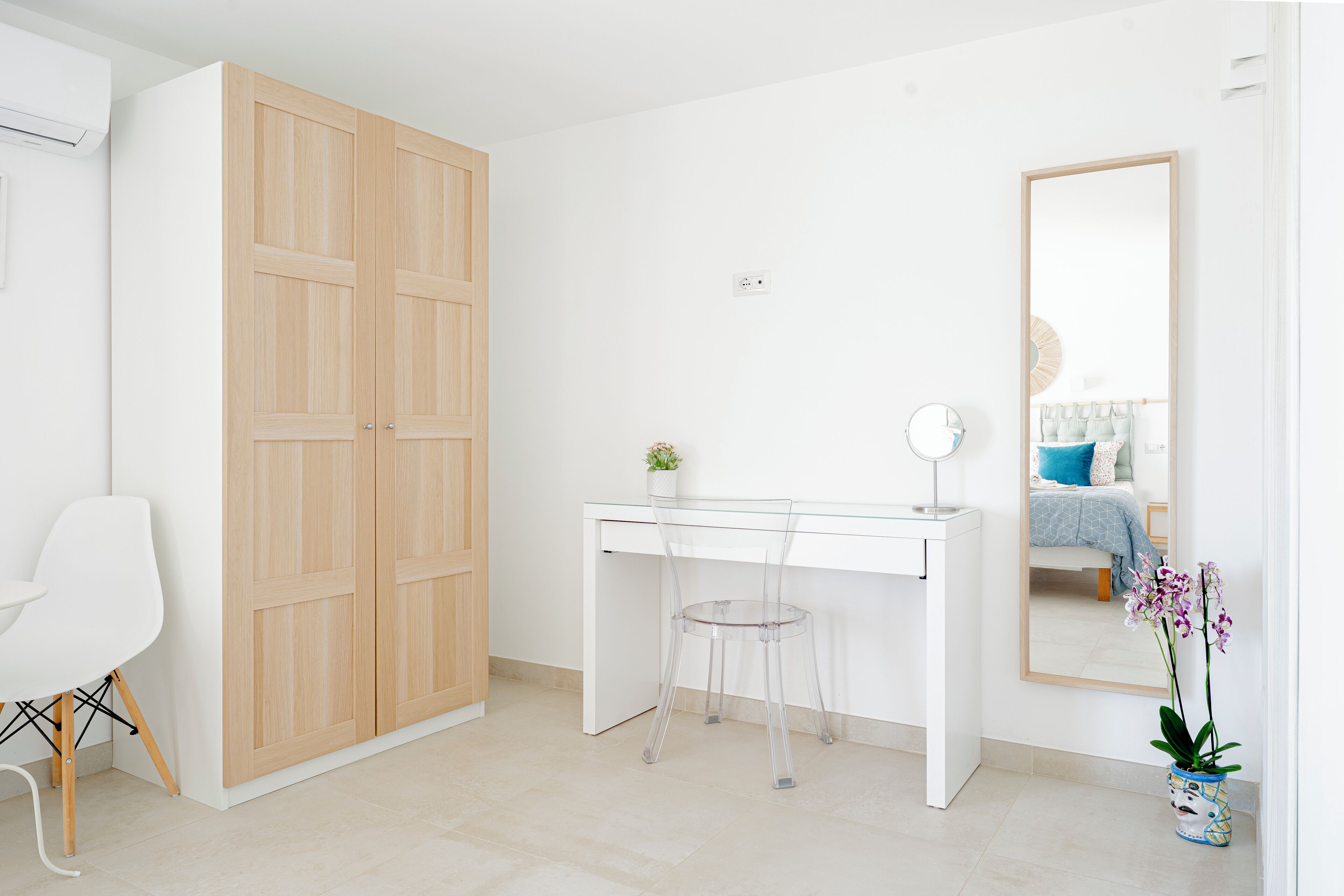 Un dormitorio con un armario de madera, un escritorio blanco con silla, una mesita de noche, un espejo y una orquídea en maceta.