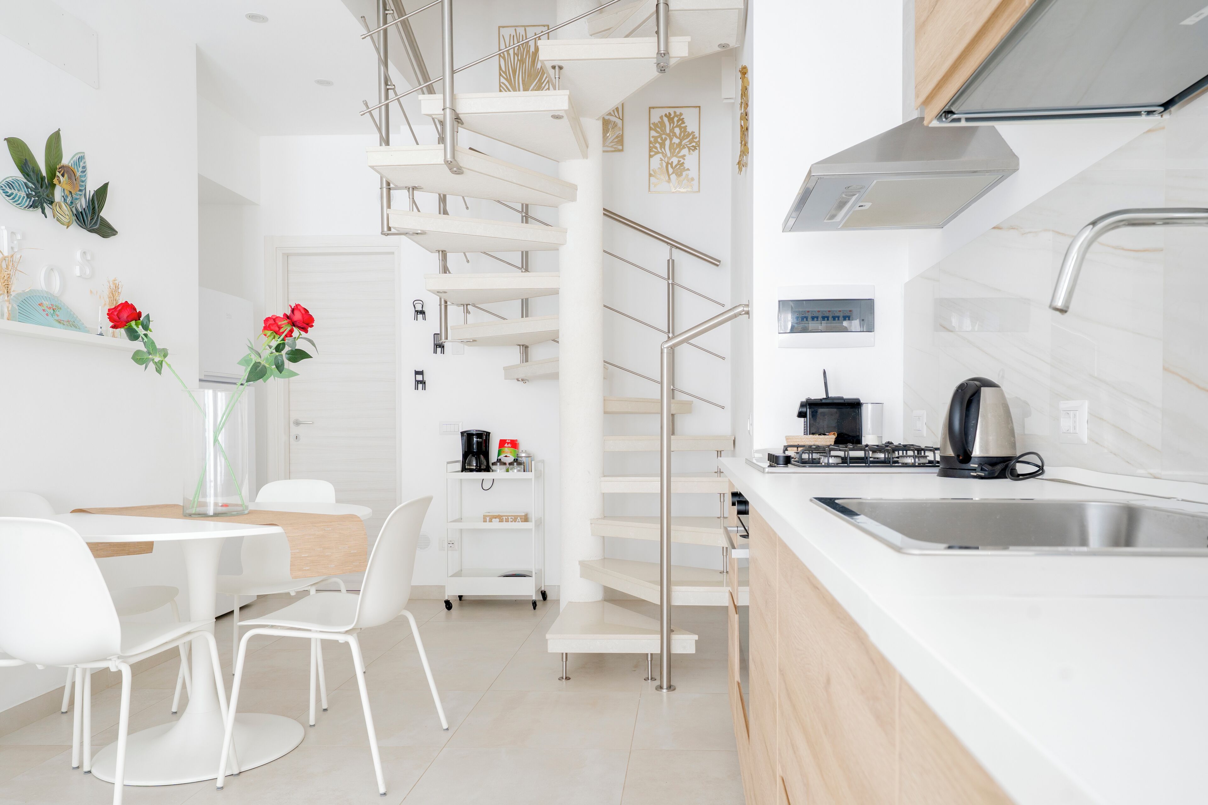 Eine moderne Küche mit weißer Arbeitsplatte, Spüle aus Edelstahl und eingebautem Herd. Ein weißer Essbereich mit Stühlen und Tisch, sowie eine weiße Treppe mit Metallgeländer.