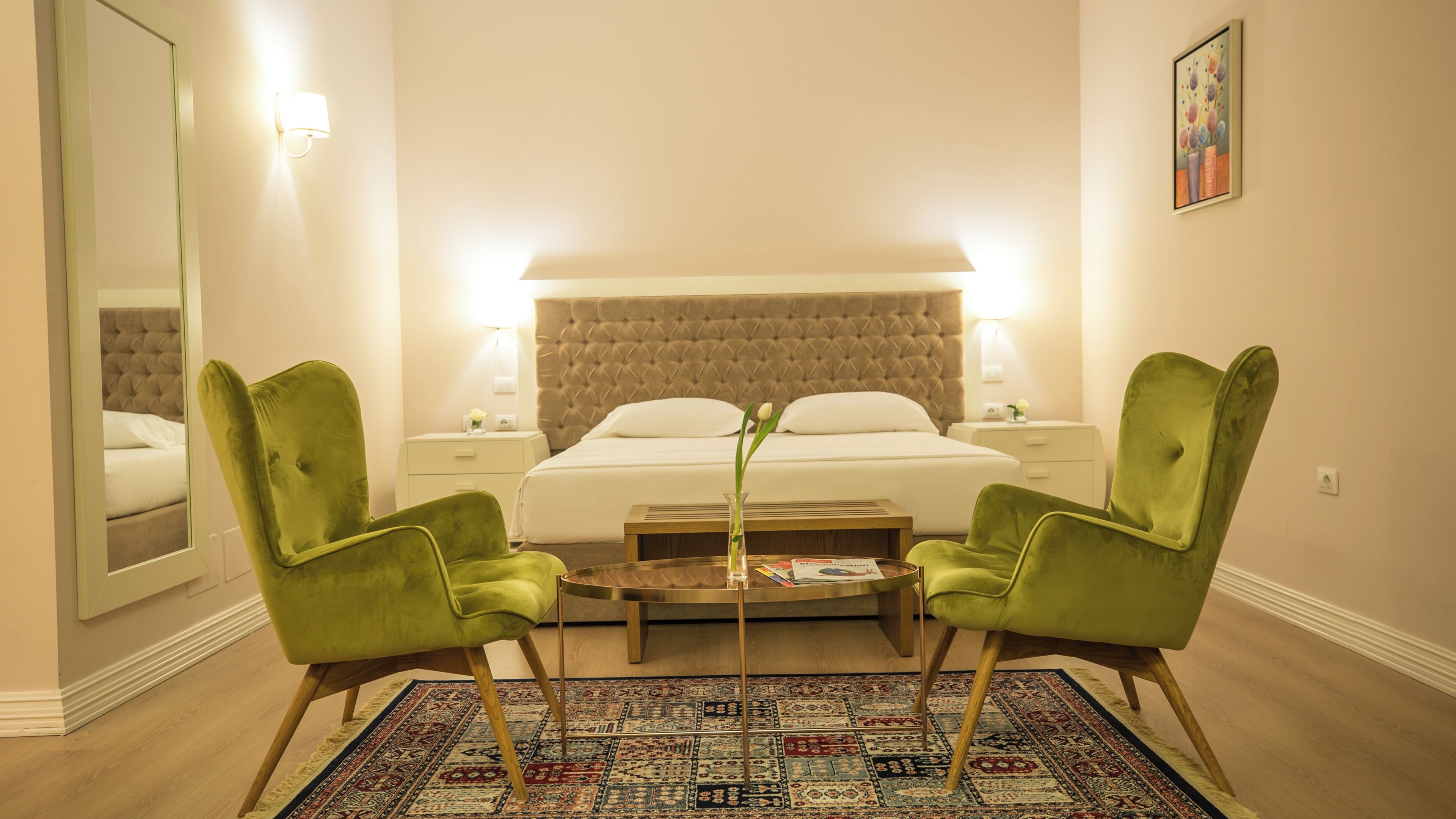 A hotel room with a large bed, two green armchairs, a coffee table, and a patterned rug.