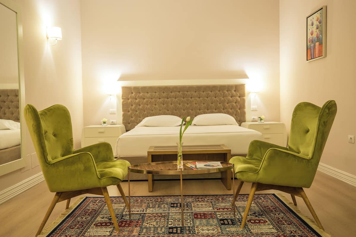 Quarto de hotel com uma cama grande, duas poltronas verdes, uma mesa de centro e um tapete estampado.