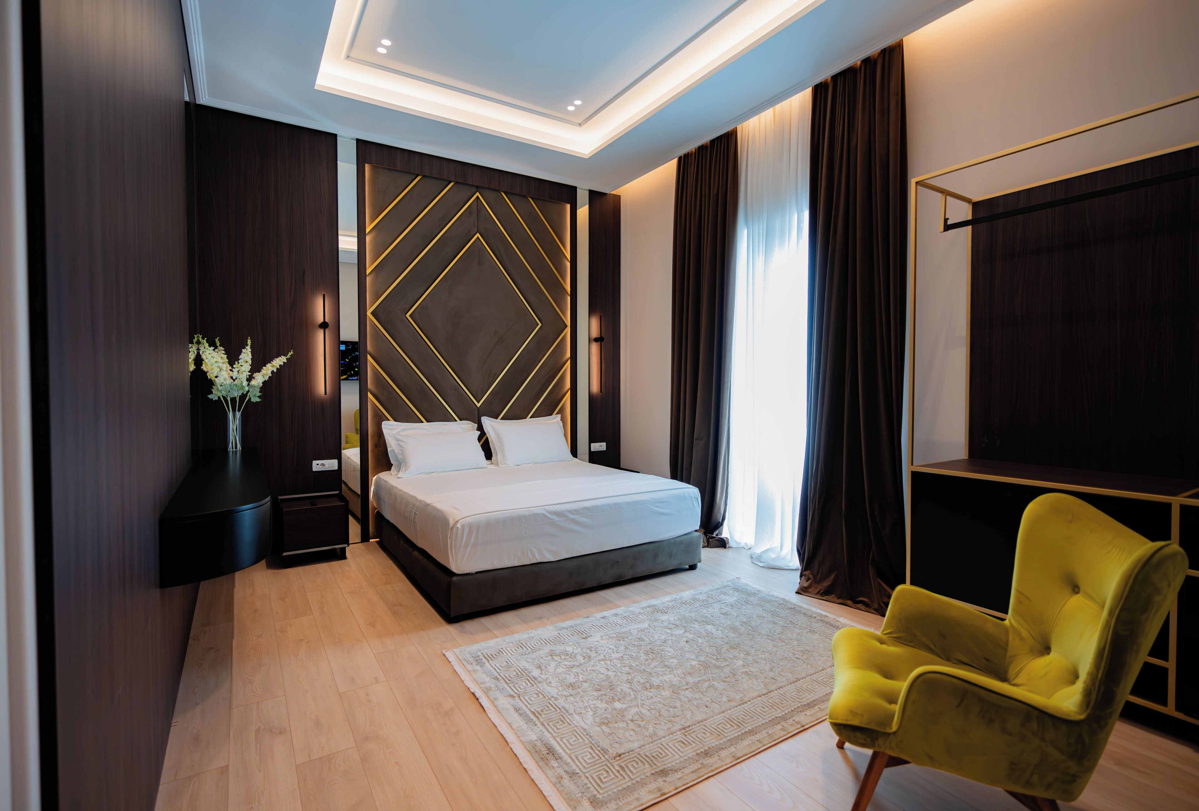 A modern bedroom with a large bed, a bedside table, a nightstand, a yellow armchair, a decorative wall panel, and a large window with curtains.