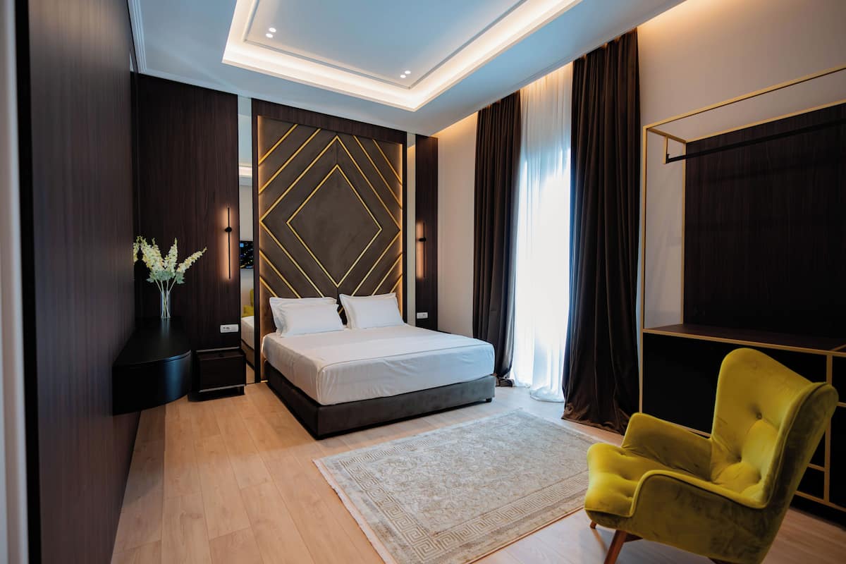 Quarto moderno com cama grande, mesa de cabeceira, poltrona amarela, painel decorativo e janela ampla com cortinas.