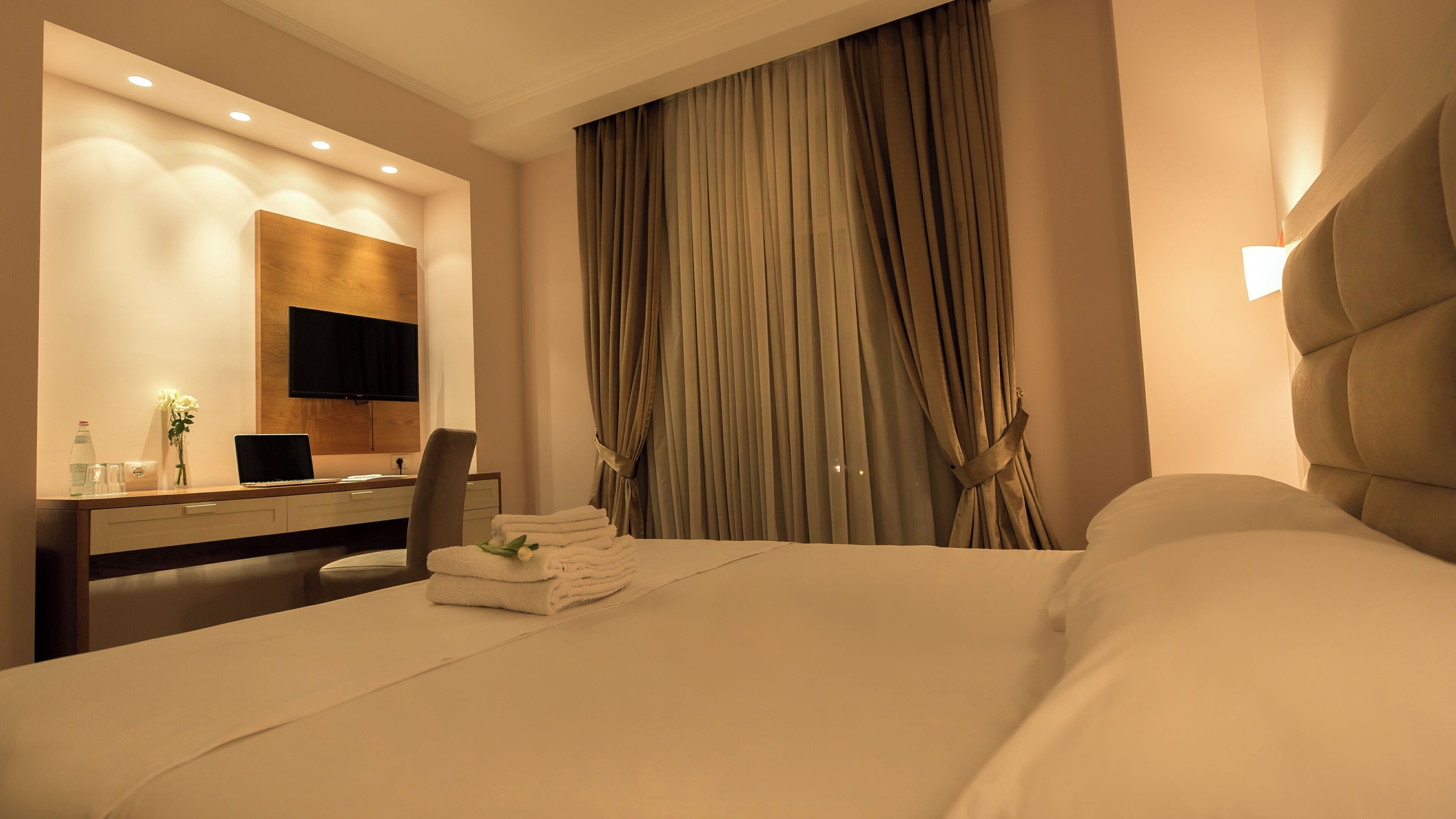 A hotel room with a bed, a desk with a computer, a chair, a TV mounted on the wall, and a large window with curtains.