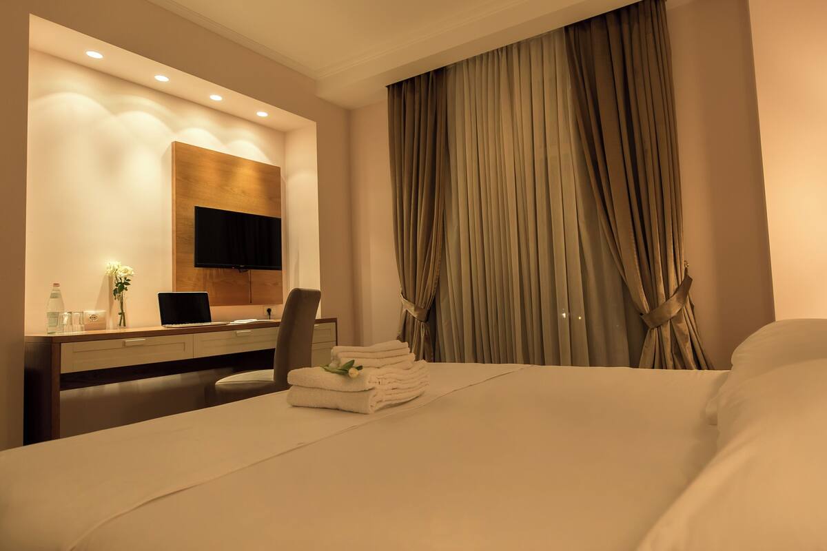 A hotel room with a bed, a desk with a computer, a chair, a TV mounted on the wall, and a large window with curtains.