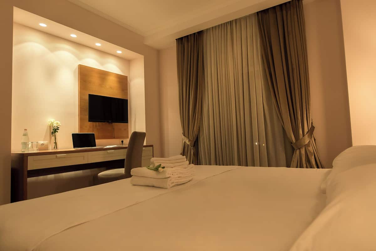 Quarto de hotel com cama, escrivaninha com computador, cadeira, televisão presa na parede e uma janela grande com cortinas.