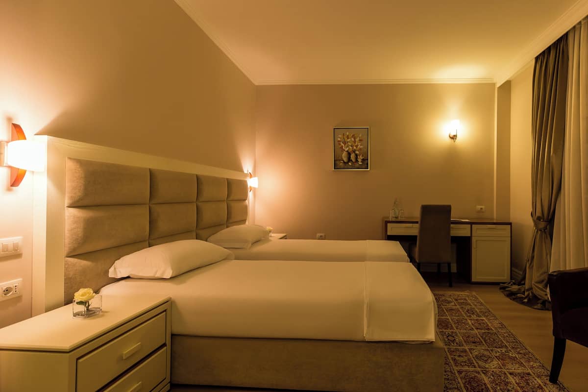 Quarto de hotel com duas camas, uma escrivaninha, uma cadeira e uma janela grande com cortinas.