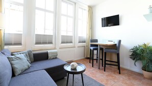 Ein modernes Wohnzimmer mit grauem Sofa, einem kleinen runden Tisch, einem an der Wand befestigten Fernseher und einer Topfpflanze.