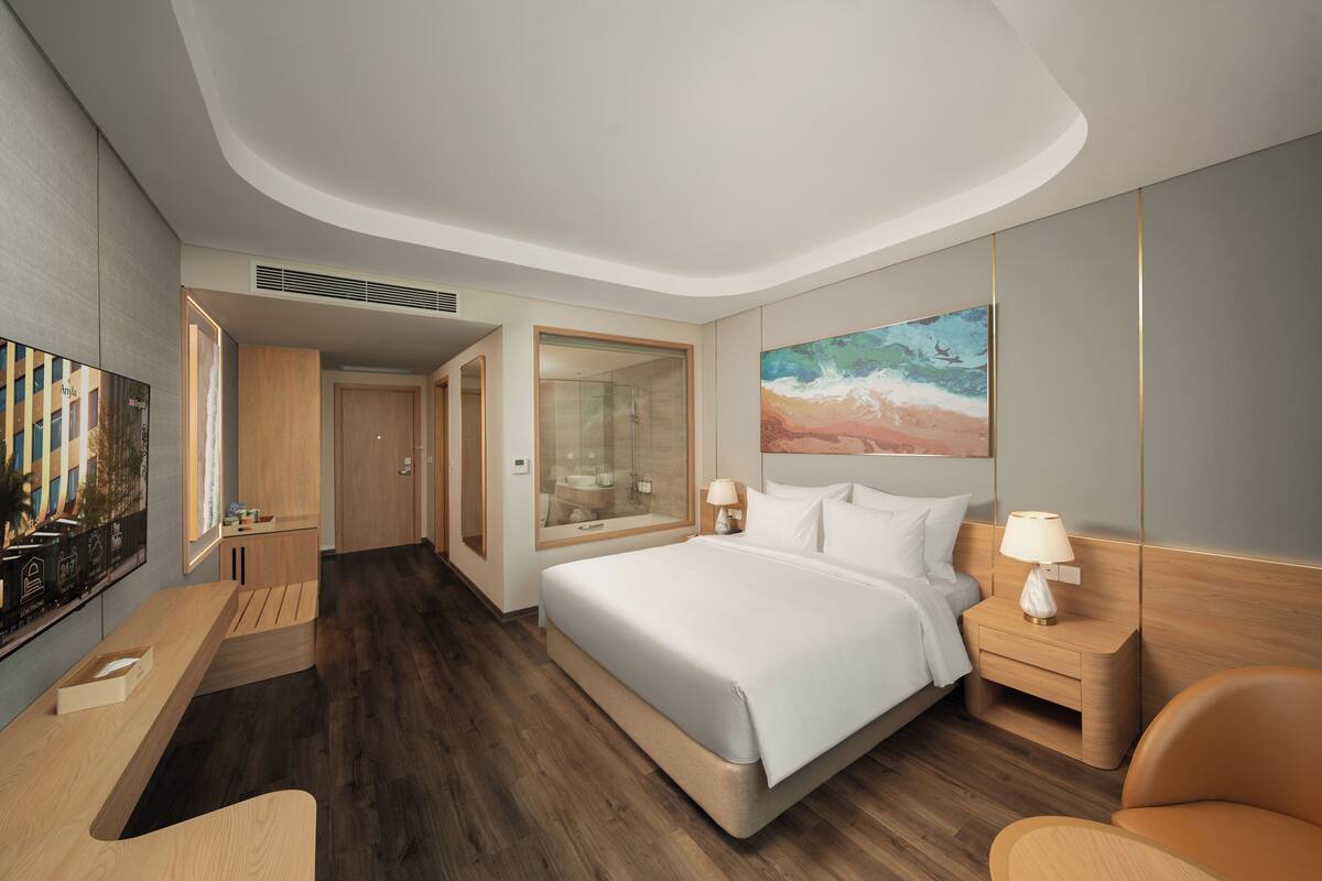 Une chambre d’hôtel moderne, dotée d’un grand lit, d’un bureau, d’une chaise, d’une télévision et d’une salle de bain visible à travers une porte en verre.