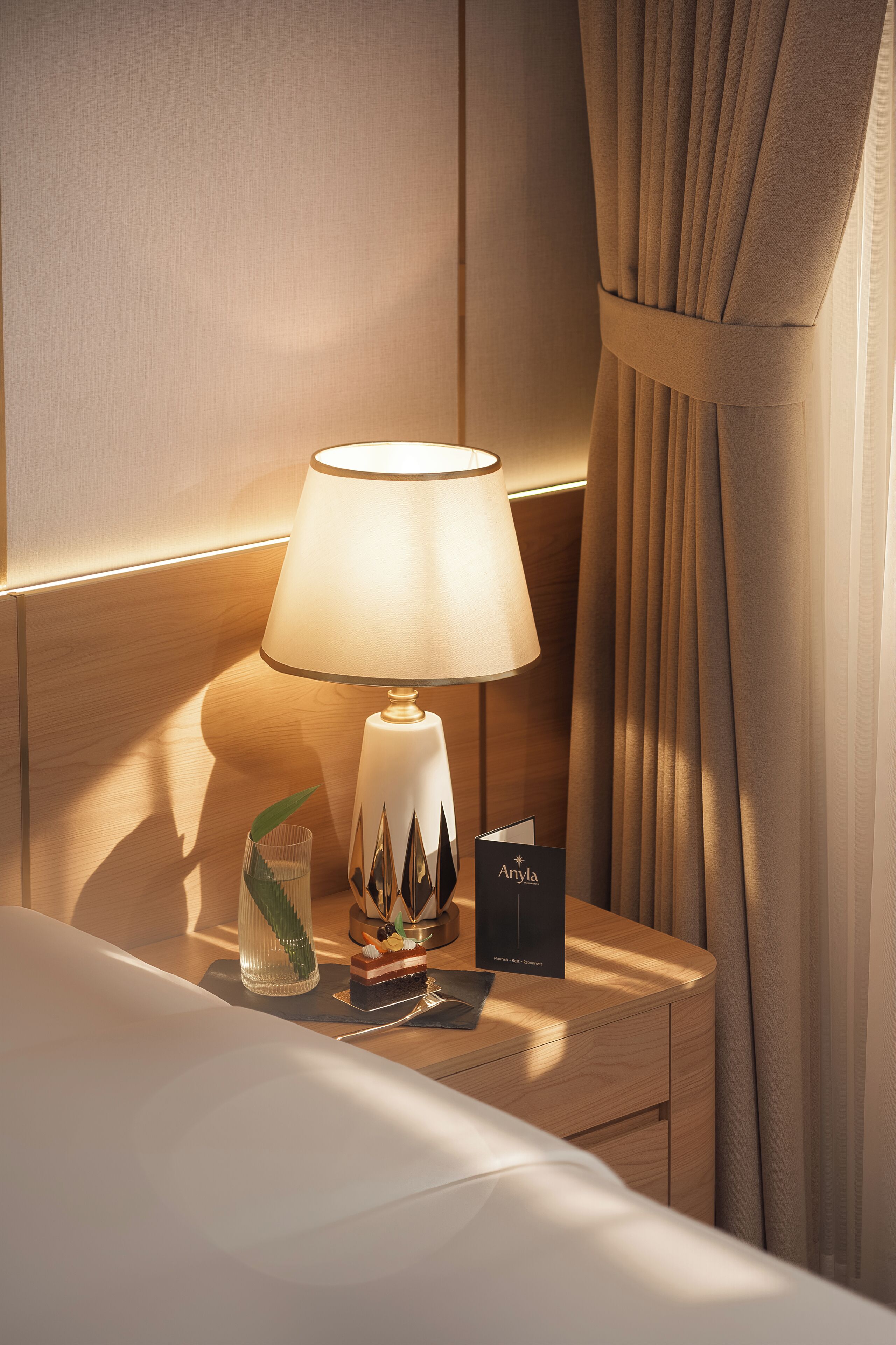 Een net opgemaakt bed met een witte deken, een bedlampje, een klein decoratief vaasje, en een naambordje op een houten nachtkastje.