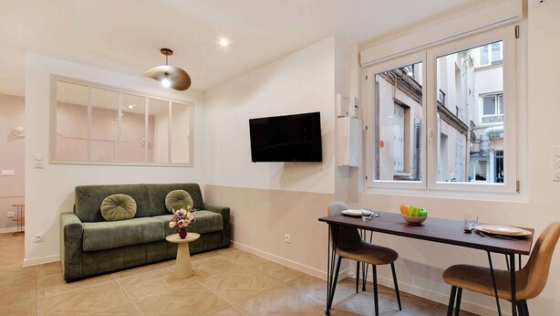 Een moderne woonkamer met een groene bank, een eettafel met stoelen, een aan de muur bevestigde televisie en een raam met uitzicht op de stad.
