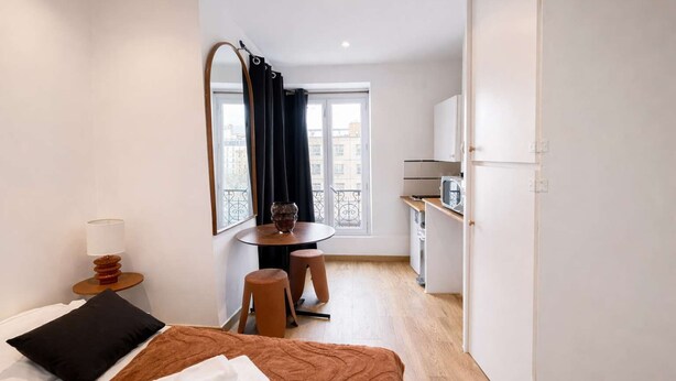 Une chambre d’hôtel compacte comprenant un lit, une petite table ronde avec deux tabourets, un coin cuisine et une grande fenêtre avec des rideaux.
