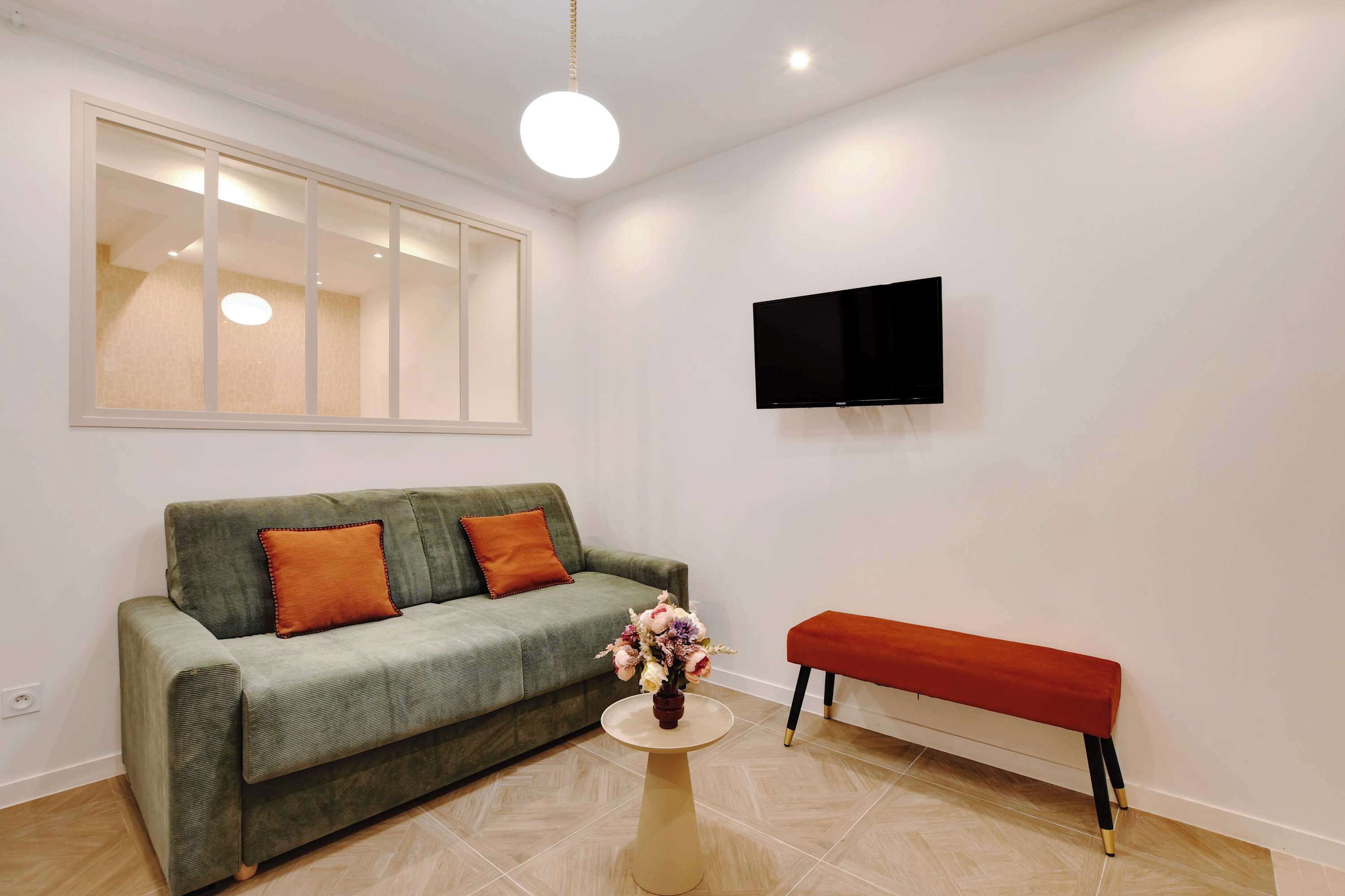 Ein modernes Wohnzimmer mit einem grünen Sofa, einer roten Bank, einem runden Couchtisch und einem an der Wand montierten Fernseher.