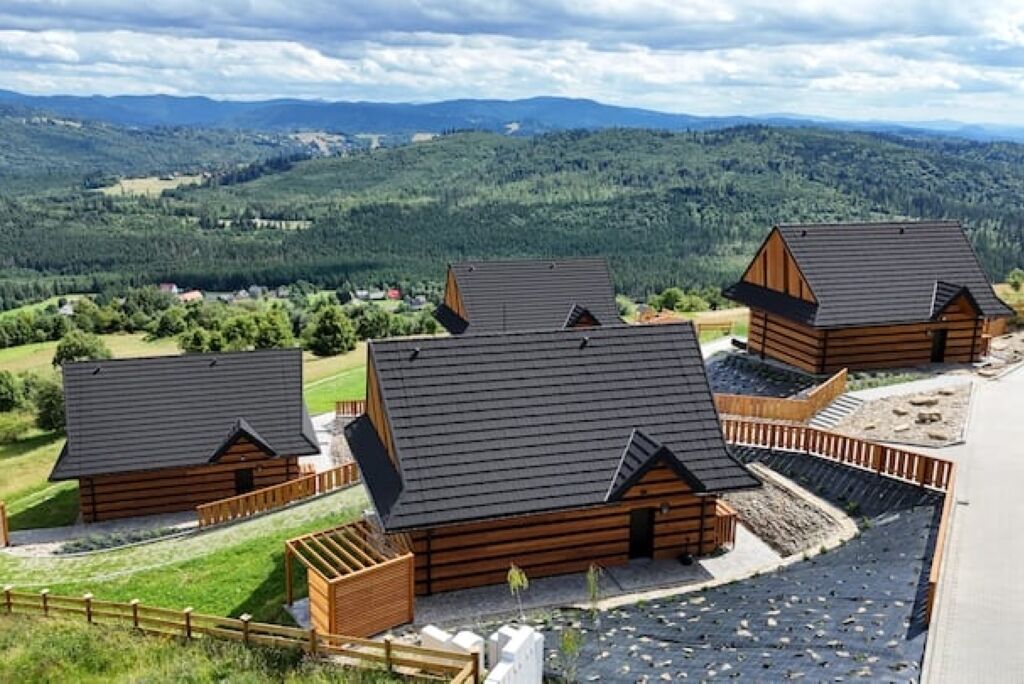 Une rangée de chalets en rondins de bois, avec des toits inclinés, entourés d’une clôture en bois, se dressant sur un fond de collines verdoyantes et d’un ciel nuageux.