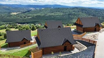 Une rangée de chalets en rondins de bois, avec des toits inclinés, entourés d’une clôture en bois, se dressant sur un fond de collines verdoyantes et d’un ciel nuageux.