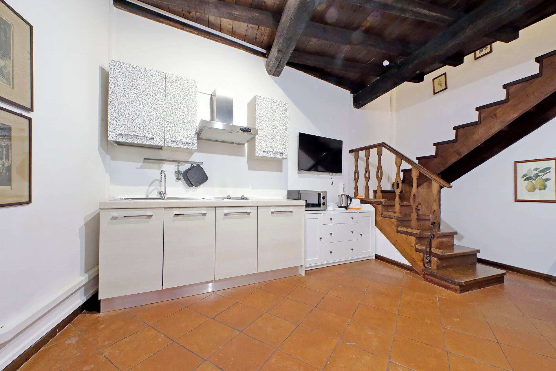 Una cocina con gabinetes blancos, piso de baldosas, una escalera de madera y un televisor en la pared.