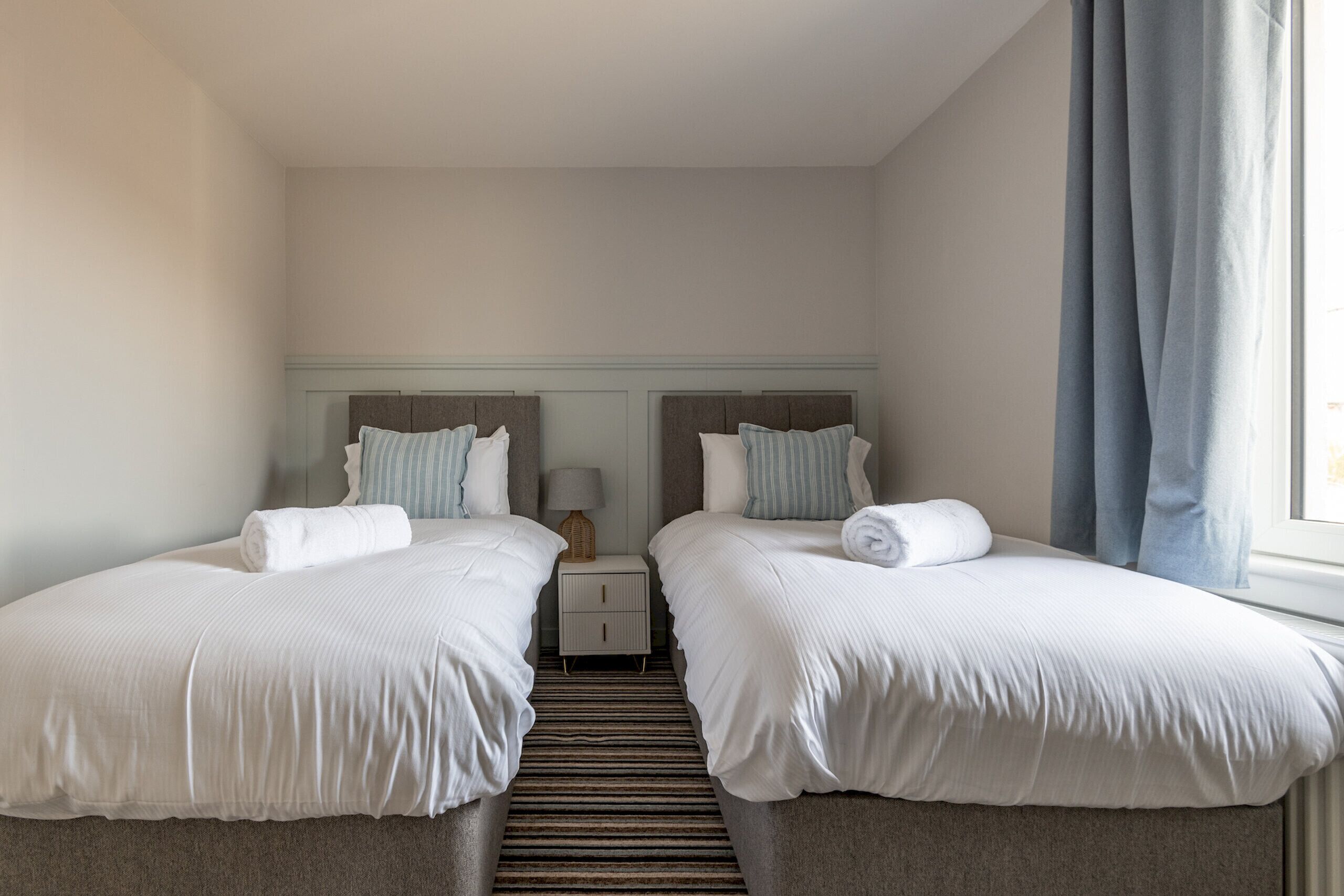 A twin room with two beds, a nightstand, a lamp, and a window with curtains.